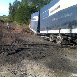 Ciężka ciężarówka dostawcza utknęła na błotnistej i niedokończonej drodze wiejskiej w budowie, a w oddali osoba oceniająca sytuację oczekuje na pomoc drogową.