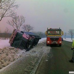 Na poboczu zaśnieżonej drogi leży przewrócona ciężarówka. Na miejscu jest pomoc drogowa.