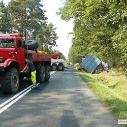 Ekipa pomocy drogowej we Wrocławiu z lawetą przygotowuje się do usunięcia przewróconej niebieskiej przyczepy z pobocza wysadzanej drzewami drogi.