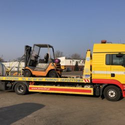 Żółta ciężarówka pomocy drogowej z przyczepą platformową przewożącą pomarańczowy wózek widłowy, zaparkowana na przestronnym obszarze przemysłowym lub usługowym pod czystym, błękitnym niebem.