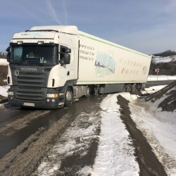 Półciężarówka z białą kabiną i zieloną przyczepą porusza się po wyboistym kawałku drogi pokrytym śniegiem i widocznymi znakami robót drogowych, wskazującymi trudne warunki jazdy w pobliżu Trzebnicy.