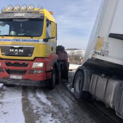 Czerwono-żółta laweta pomocy drogowej „Jarex” we Wrocławiu przygotowująca się do udzielenia pomocy na zaśnieżonym poboczu, na pierwszym planie widoczna duża biała ciężarówka.