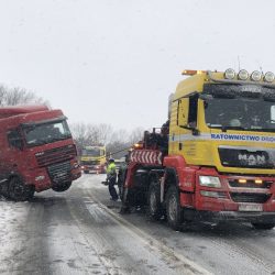 Zaśnieżona scena pomocy drogowej we Wrocławiu, gdzie jasnożółty laweta przygotowuje się do ciągnięcia czerwonej półciężarówki pod obfitymi opadami śniegu.