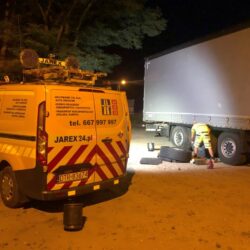 Pomoc drogowa w akcji w nocy, z technikiem pracującym przy kole ciężarówki w pobliżu samochodu serwisowego wyposażonego w światła ostrzegawcze i sprzęt komunikacyjny.