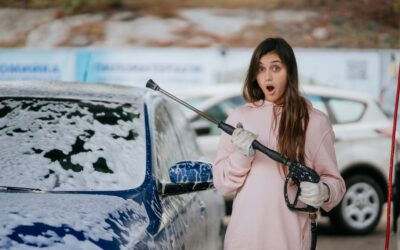Młoda kobieta zaskoczona, trzymając myjkę ciśnieniową do samochodu zimą, na tle samochodu pokrytego pianą.