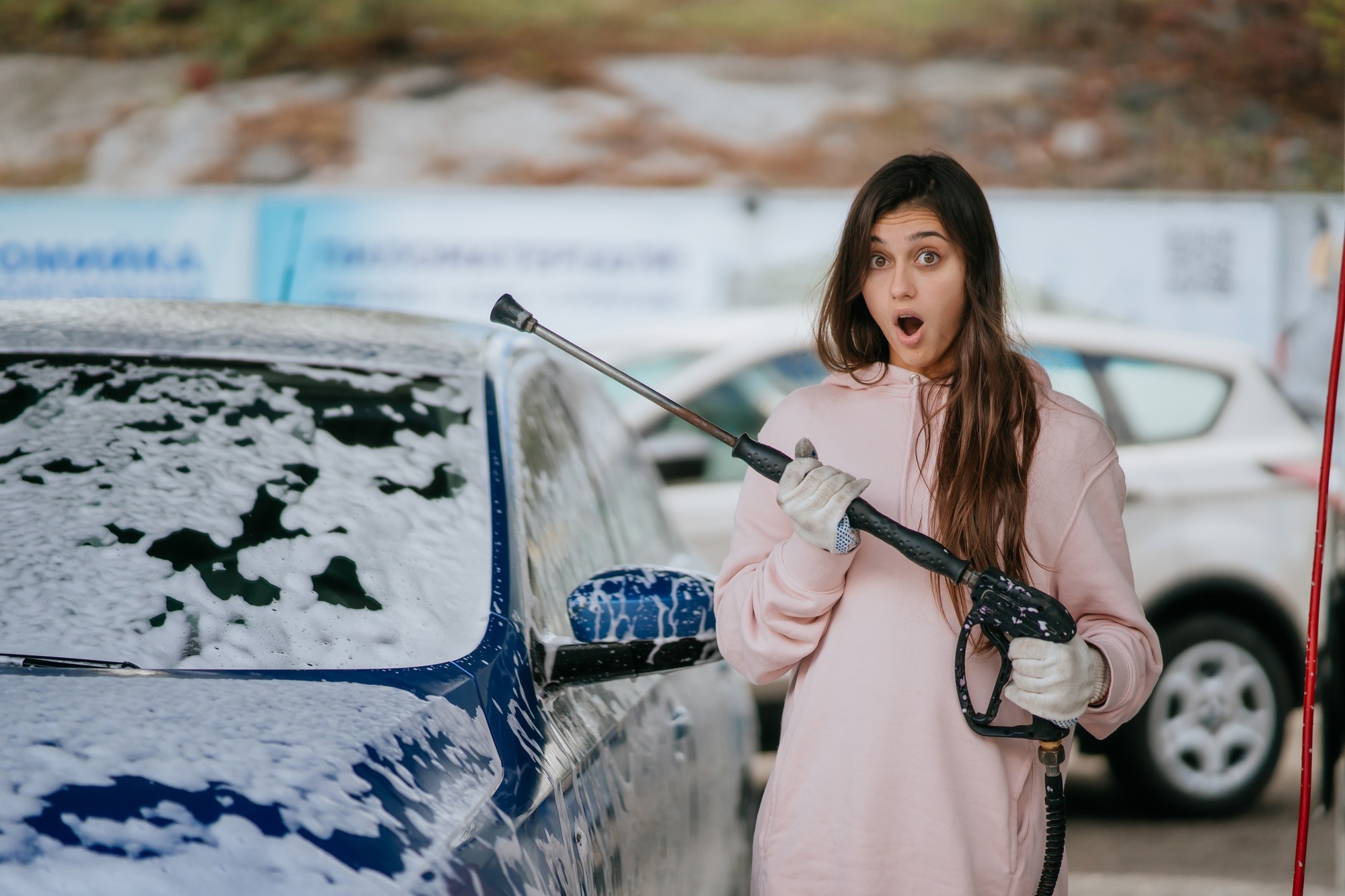 Młoda kobieta zaskoczona, trzymając myjkę ciśnieniową do samochodu zimą, na tle samochodu pokrytego pianą.
